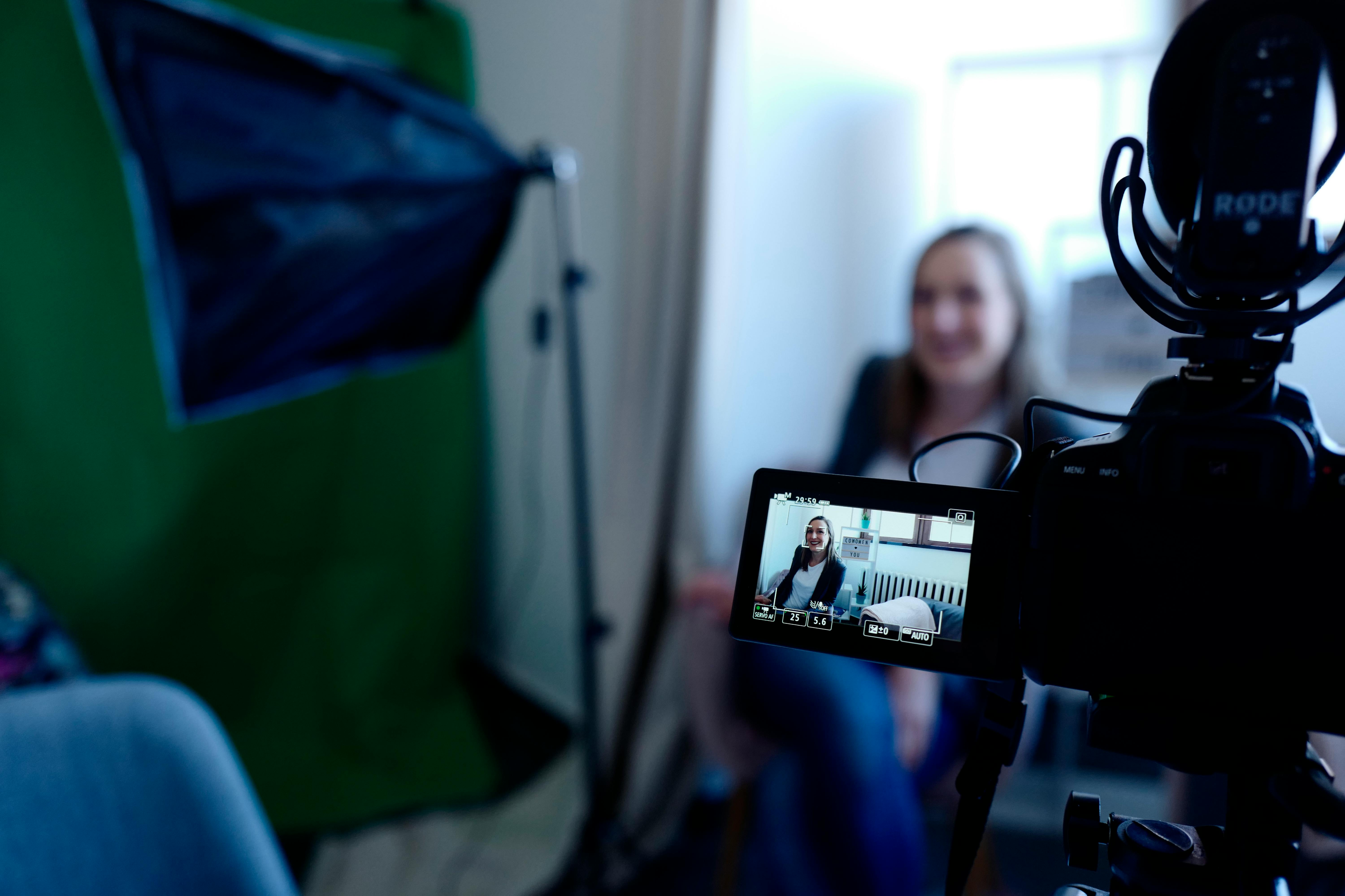 A woman sitting in front of a camera for an interview. Shown through a camera viewfinder. Next to her is lighting equipment and a green screen.